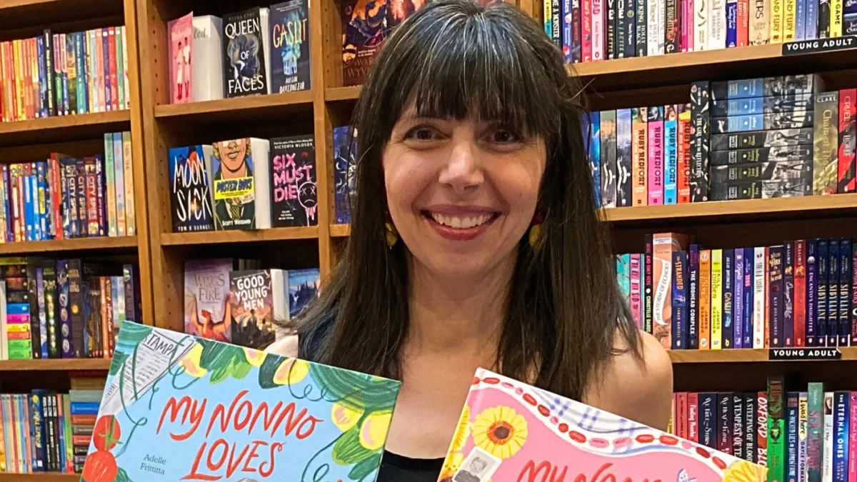 A woman is standing in front of a book shelf smiling. She is holding 2 books.
