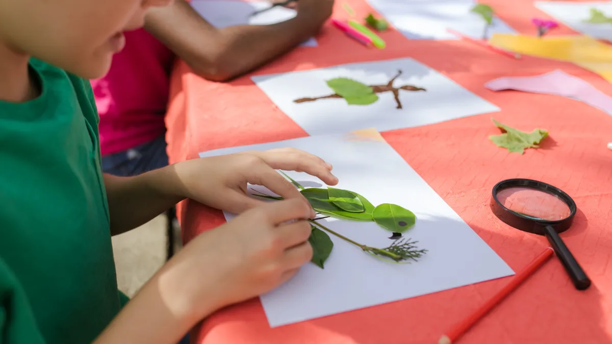 A child is using leaves and sticks to create a bird image on white paper. There is a magnifying glass and pencil nearby