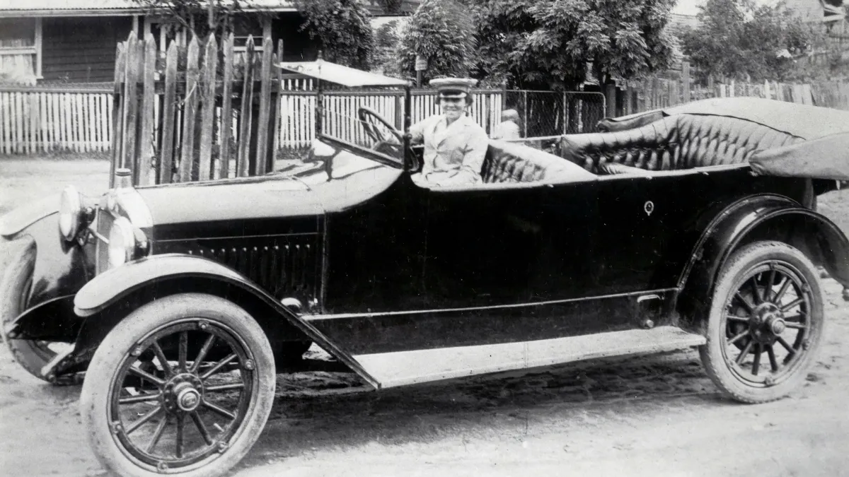 Black and white photograph of a women in an old-fashioned car with no roof. The women is wearing a jacket and cap and smiling into the camera.