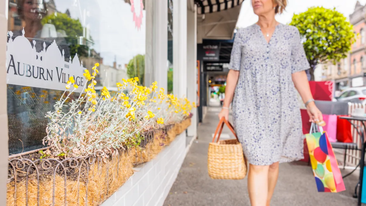 A person with shopping bags walks past a shop window lined with planter boxes containing yellow flowers