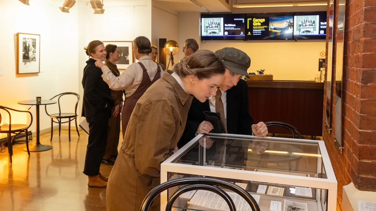 Two women peer into an exhibition cabinet in the foreground. A group of figures are chatting amiably in the background.