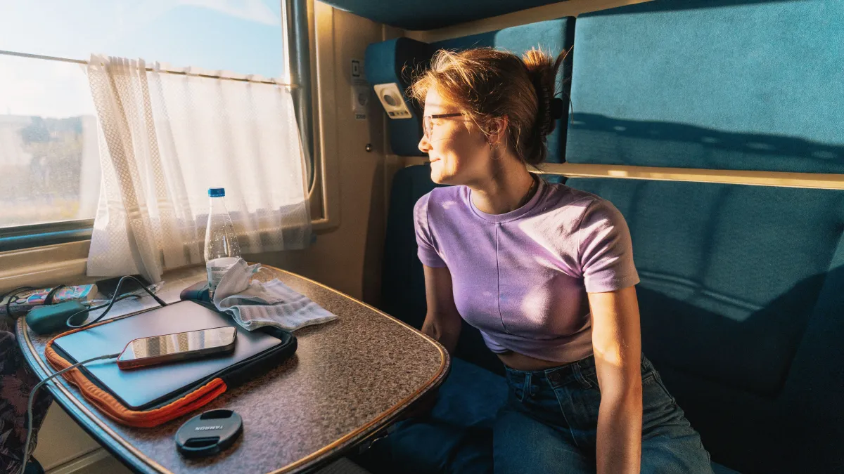 Young woman gazes out train window. On the table in front of her is a water bottle, phone, laptop and battery charger.