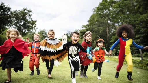 Children in colourful Halloween costumes running across a grassy park during an outdoor community event.