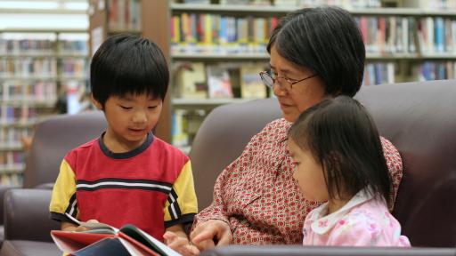 A young child reads to a grandmother and sibling