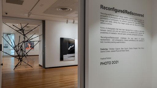 Looking into a gallery space. Wall text displays blurb about the exhibition. An artwork made of camera tripods fills a doorway, blocking the path.