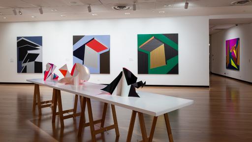 Installation view of 'Light Gestures' at Town Hall Gallery. Abstract geometric painting on the wall that are reminiscent of buildings or structures. Abstract sculptures on a table, made up of large flat surfaces and lots of angles.