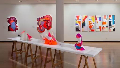 Installation view of 'Light Gestures' at Town Hall Gallery. Abstract geometric painting on the wall that are reminiscent of buildings or structures. Abstract sculptures on a table, made up of large flat surfaces and lots of angles.