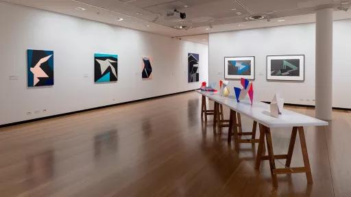 Installation view of 'Light Gestures' at Town Hall Gallery. Abstract geometric painting on the wall that are reminiscent of buildings or structures. Abstract sculptures on a table, made up of large flat surfaces and lots of angles.
