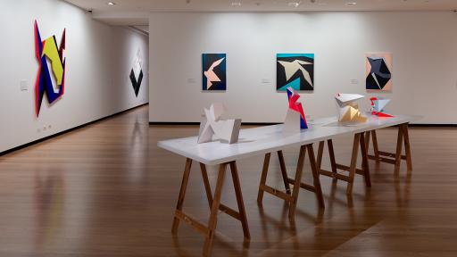 Installation view of 'Light Gestures' at Town Hall Gallery. Abstract geometric painting on the wall that are reminiscent of buildings or structures. Abstract sculptures on a table, made up of large flat surfaces and lots of angles.