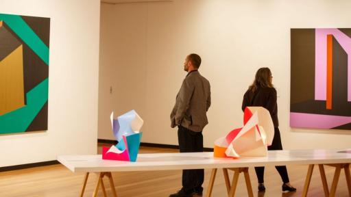Visitors to 'Light Gestures' looking at paintings on the walls. In the foreground is a table with abstract sculptures. 