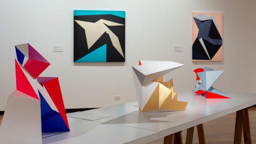 Installation view of 'Light Gestures' at Town Hall Gallery. Abstract geometric painting on the wall that are reminiscent of scrunched paper. Abstract sculptures on a table, made up of large flat surfaces and lots of angles.