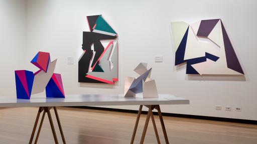 Installation view of 'Light Gestures' at Town Hall Gallery. Abstract geometric paintings on the wall made from cut out shapes. Abstract sculptures on a table, made up of large flat surfaces and lots of angles.