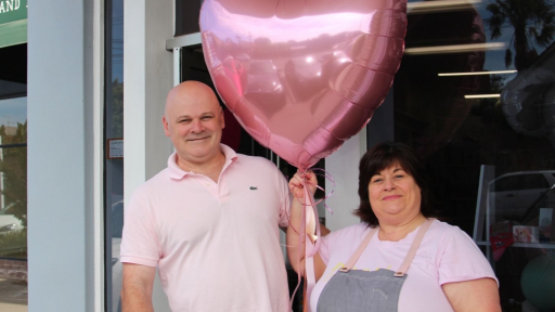 Two business owners stand outside of their business holding a giant pink balloon