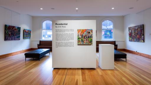 a section of movable wall in the centre of the exhibition space. On it is a blurb about the exhibition and a painting of the exterior of a house. Behind the wall are seats, windows and more paintings on the other walls.