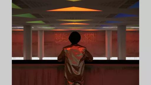A figure with their back to us leaning on a barrier, looking out across an empty space that contains four pillars.