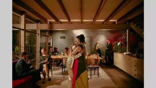 Two people hugging in the middle of a room. Behind them is a dining table with glasses and wine bottles. Around the room are people chatting to each other.