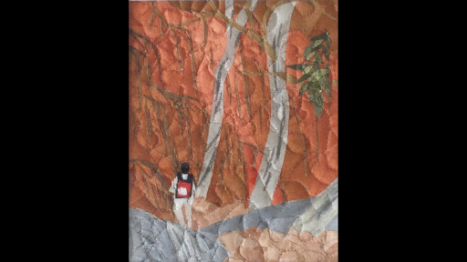 A quilted artwork of a red rock face, two tall skinny trees and a figure wearing a backpack walking.