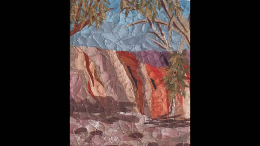 A quilted artwork of a red rock face with overhanging trees, against a blue sky.