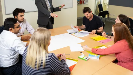 A group of people sitting at a table. One person standing, and pointing at some paper on the table.