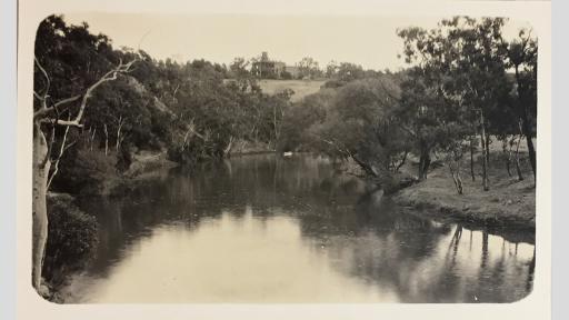 Photo of a still river framed by trees and a building on a hill in the background