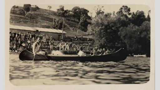 Photo of two people in a canoe on water with people on the riverbank watching in the background