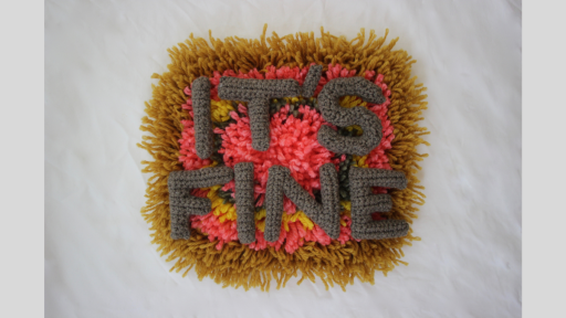 A piece of carpet with brown, pink, yellow, and grey wool in concentric cicles, overlaid with crocheted letters spelling out 'It's fine'