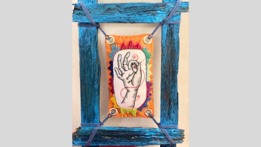 A rustic blue wooden frame with a piece of felt art suspended in the middle of the frame showing a hand holding a sewing needle with red thread