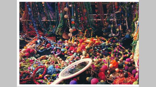 Photo by Elisabeth Frank of a table covered in beads on necklace strongs in piles, with a hand mirron lying on top and more beaded necklaces hanging in the background