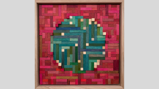 A framed piece of art made of wooden rectanges arranged to make a mainly green/blue circle surrounded by tesselating pink blocks. Some of the blocks in this piece are raised so they emerge from the artwork.