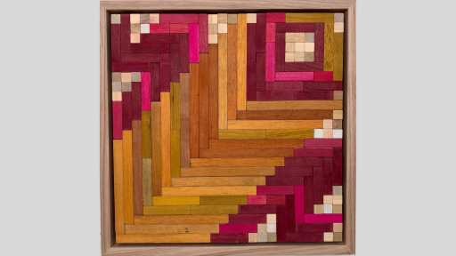 A framed piece of art made of wooden rectanges arranged in a herringbone pattern with tesselating brown blocks on a diagonal through the middle, and pink blocks with some light brown/white blocks in 2 corners and forming a square in the 3rd corner
