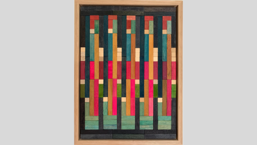 A framed piece of art made of wooden rectanges arranged in columns with bright pink, blue and some light and dark brown pieces in the middle of the columns surrounded by dark grey pieces