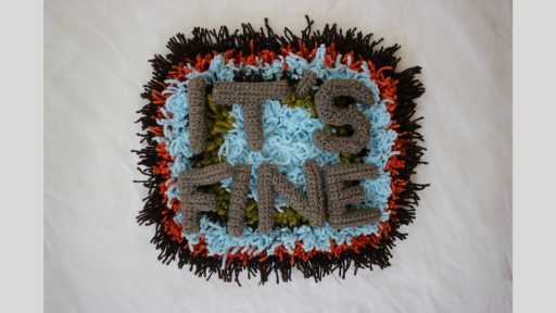A piece of carpet with black, red, light blue, and green wool in concentric circles, overlaid with crocheted letters spelling out 'It's fine'