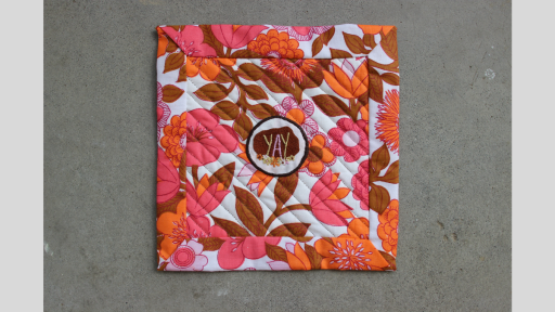 A square quilt using orange and pink flowered material, that has a patch in the middle that says 'Yay' and is decorated with embroidered flowers