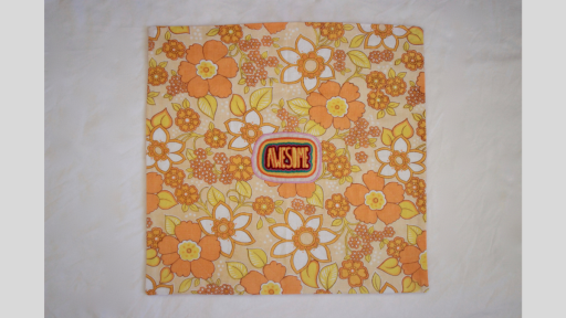 A square quilt using orange and yellow flowered material, that has a patch in the middle that says 'Awesome' and is surrounded by a circular rainbow to the edge of the patch