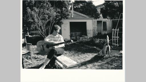 Photo by Elisabeth Frank of a person sitting in a backyard holding a guitar while a dog stretches alongside them