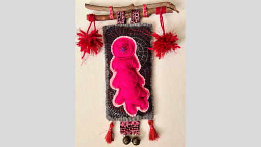 A piece of artwork on felt of a pink tardigrade. The artwork is hanging from a branch by patterned material, and there are pompoms hanging off the branch and a piece of materials with 2 bells hanging under the felt piece.
