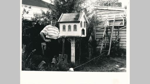 Photo of a person in a backyard leaning against a guitar and standing in front of a large birdhouse shaped line a house with windows and a door