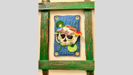 A rustic green wooden frame with a piece of felt art in the middle of the frame showing a skull with mushrooms growing out of the skull which are raised from the felt so they emerge from the piece.