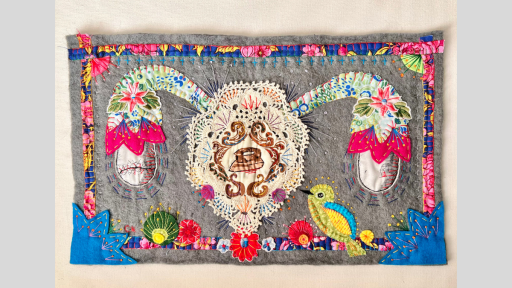 A piece of artwork on material of anatomically correct overies made out of material with a doily in the centre that has the image of a iron on it. The felt is bordered by a bright material fram with birds and flowers on it.
