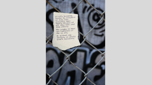 A piece of torn paper with a poem printed from a typewriter, stuck to a chainlink fence with a grey wall behind it that is graffitied with black spray paint
