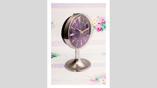 A silver metal clock with a purple face showing 10:15, in front of a wallpaper background with pink flowers