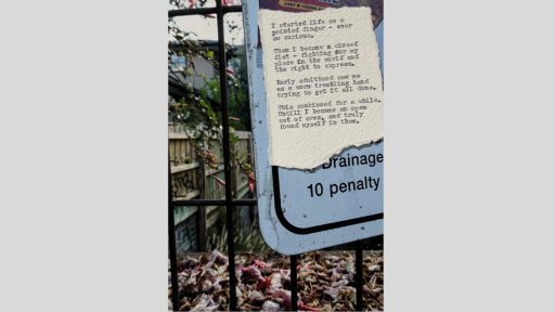 A piece of torn paper with a short piece of text printed from a typewriter, stuck to a sign on a fence outside with a backyard fence and greenery behind it