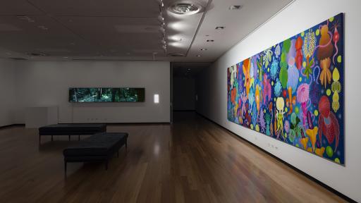The exhibition space for Above the Canopy, showing a large painting covering most of a side wall with bright multicolour flowers on a blue canvas, and a screen on the far wall showing another artwork