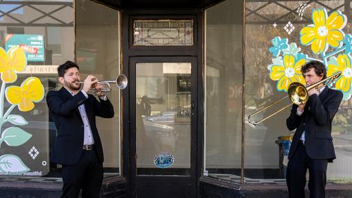 Two trumpeters playing their trumpets in front of an empty storefront window that has bright yellow floral artwork painted on it