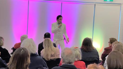 An Elvis tribute performer singing in a white jumpsuit in front of a seated crowd