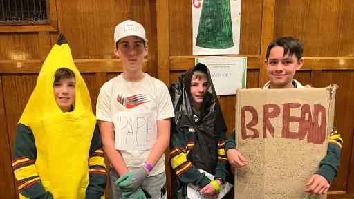 Four children standing behind a table. One is in a banana costume, one is wearing a black plastic bag and two are holding signs saying paper and bread.