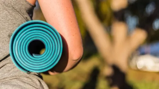 Rear view of person carrying a rolled-up green yoga mat under their arm. Only the arm and part of the torso are visible.
