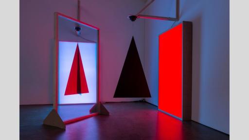 A mirror reflecting the image of a red triagle with a black line through the centre, next to a bright red lit rectange and a hanging 3D black pyramid in front of it.