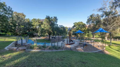 Man-made garden with a tall brown sculpture, rock feature and two blue umbrellas to the right. It includes concrete and tan bark sections and is surrounced by grass. There are trees in the background.