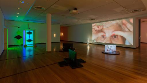 Large indoor art installation space with hardwood floors and three scattered televisions. To the right there is a photo of the top half of an elderly woman's face.